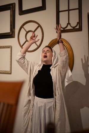 Dramatic scene with performer in black and white outfit raising arms expressively against wall decorated with frames.
