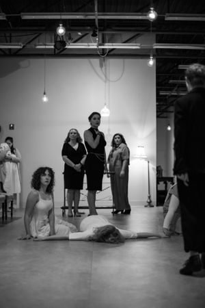 A dramatic black and white performance art scene with people in an industrial space lit by hanging light bulbs.