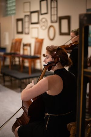 Musician plays cello in black dress against wall decorated with vintage frames.