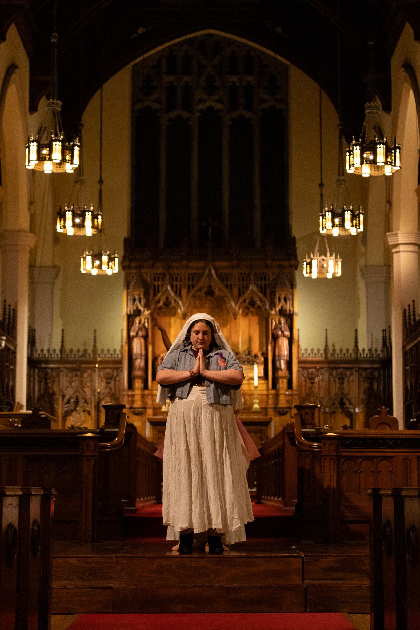 A figure stands in white clothing inside a historic gothic church sanctuary with ornate wooden details and hanging chandeliers.