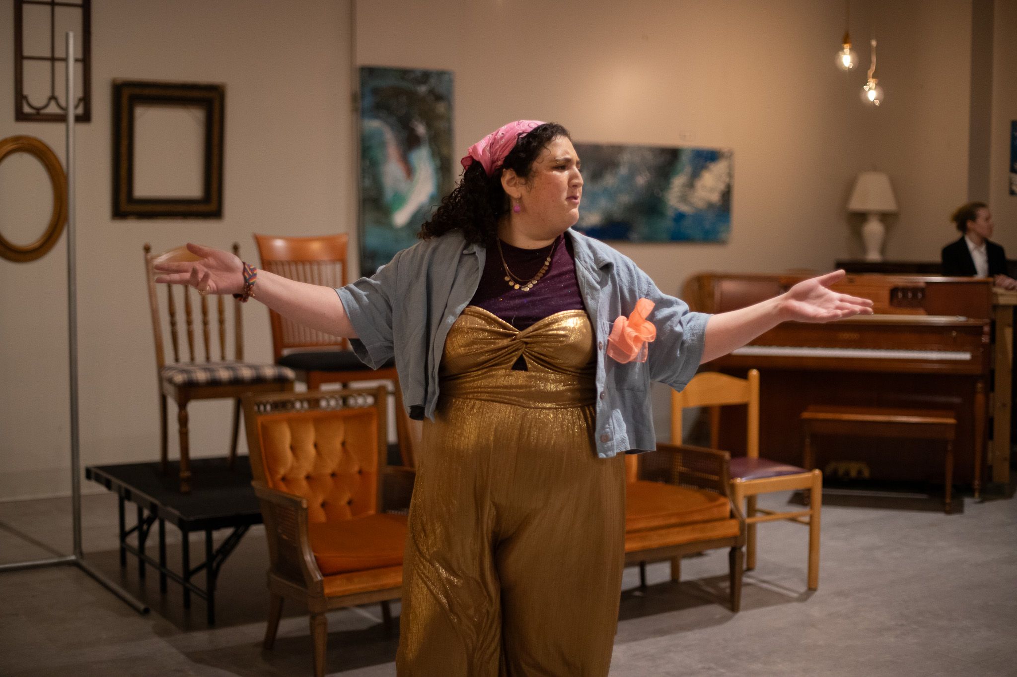 Performer in yellow overalls and denim jacket poses dramatically in a room with wooden chairs and framed art.