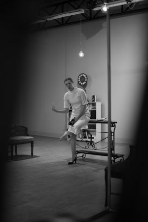 A performer in all white sits on a piece of furniture in a minimalist black and white studio setting.