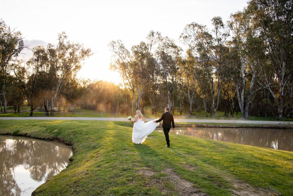 Wedding couple walks along grassy hill at sunset with eucalyptus trees in background creating romantic ambiance.