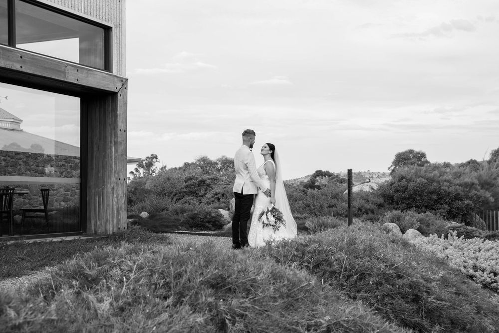 Bride and groom embrace in modern architectural setting with expansive landscape views in black and white photography.