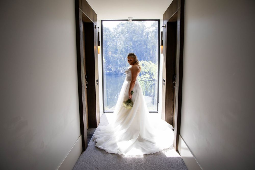 A bride in a white wedding dress stands silhouetted in a doorway holding her bouquet against a bright outdoor backdrop.