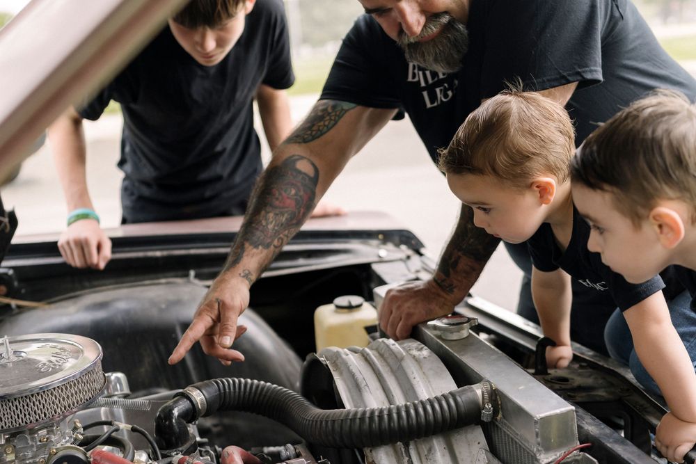Two children looking under the hood of a car while being shown engine components by an adult with tattoos.