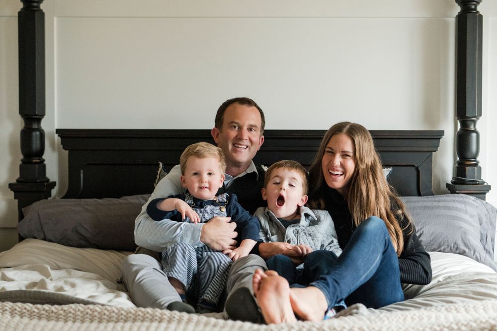 A family lounges together on a gray bed, sharing joyful laughter and embraces against a white wall backdrop.