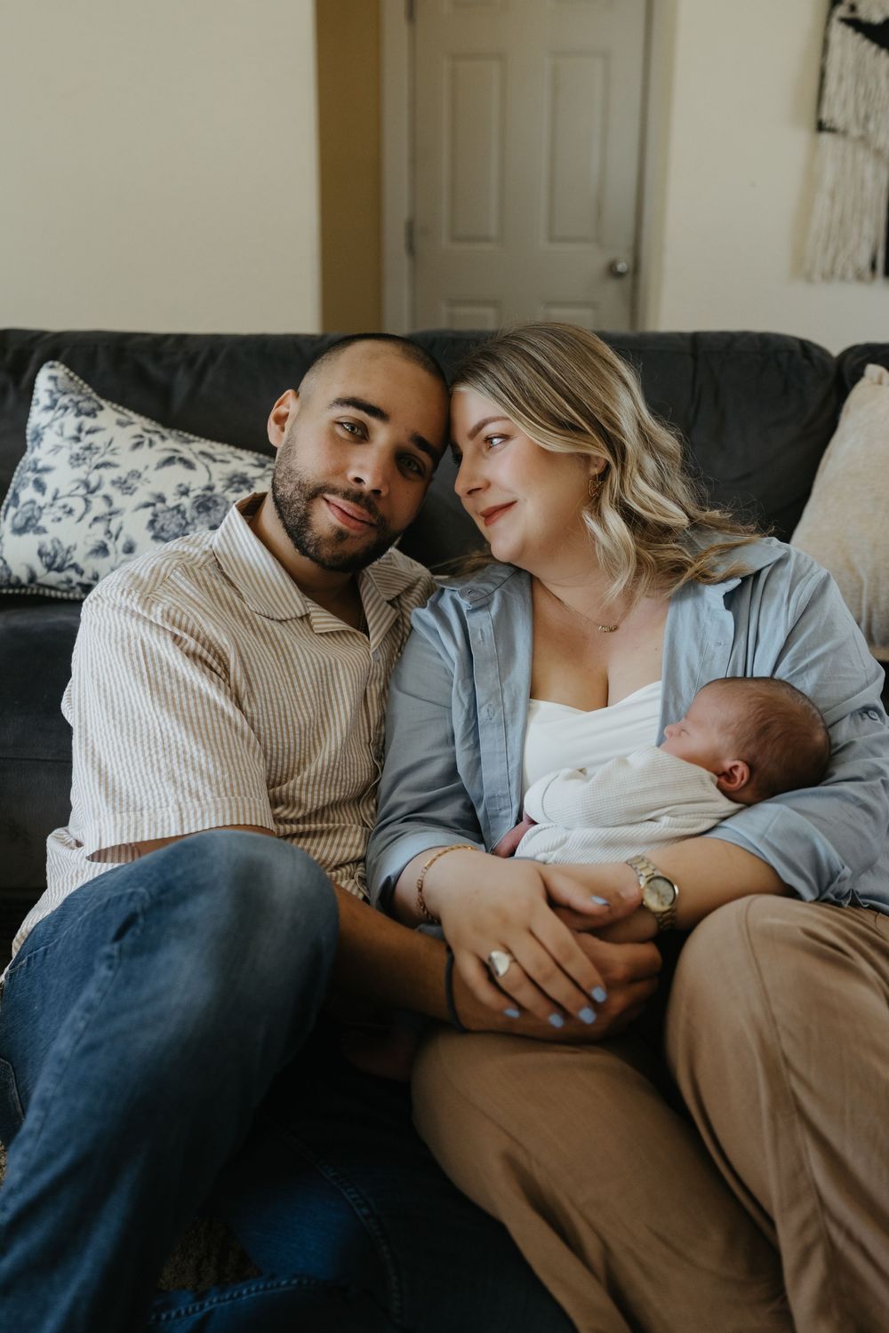 New parents sitting on a couch together, smiling, as the mother holds their sleeping newborn baby.