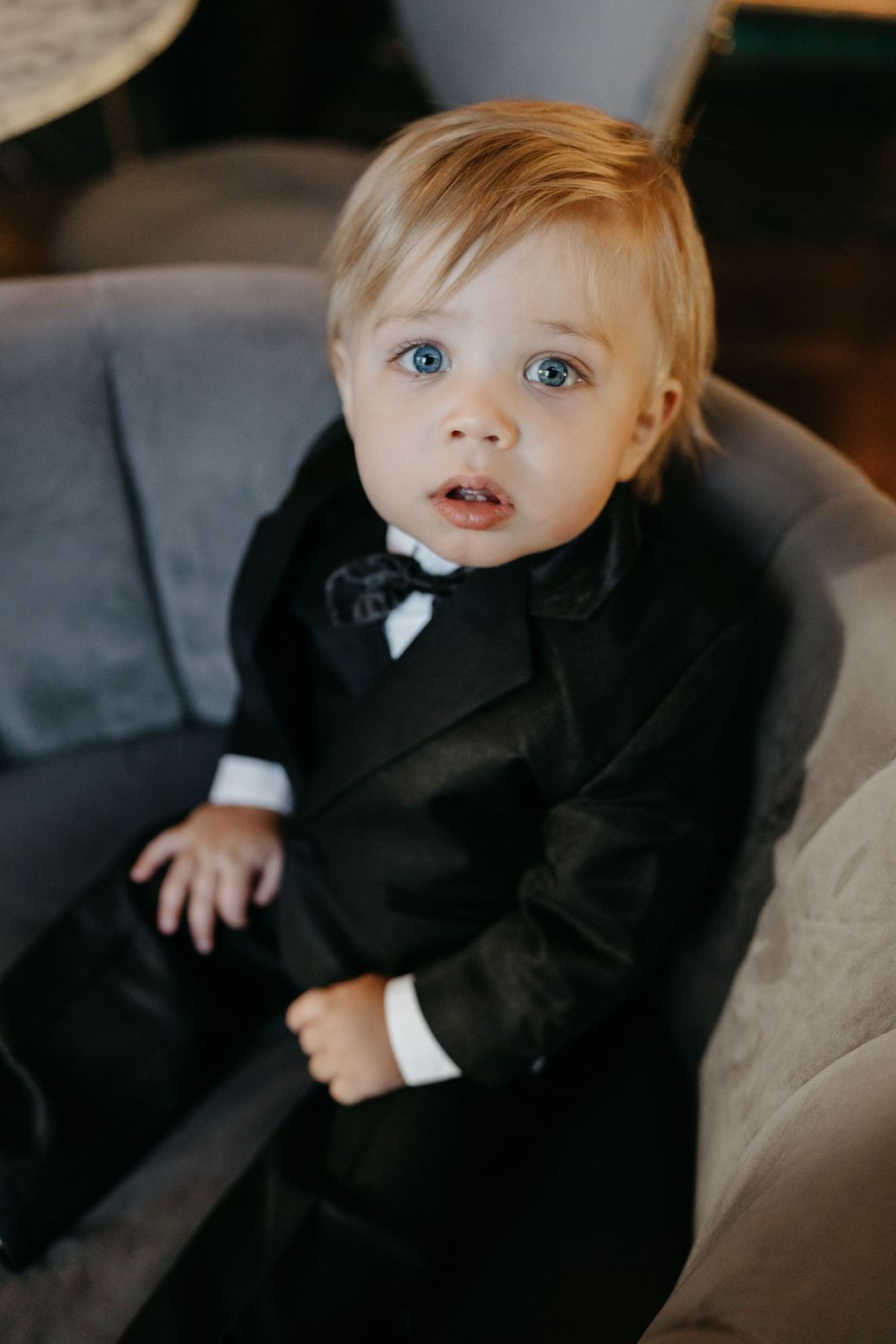 A very young child dressed in a formal black tuxedo and bow tie sits on a gray couch.