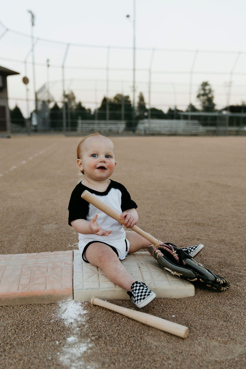 Small child sits on baseball field with baseball bat and glove wearing black and white outfit and checkered shoes.