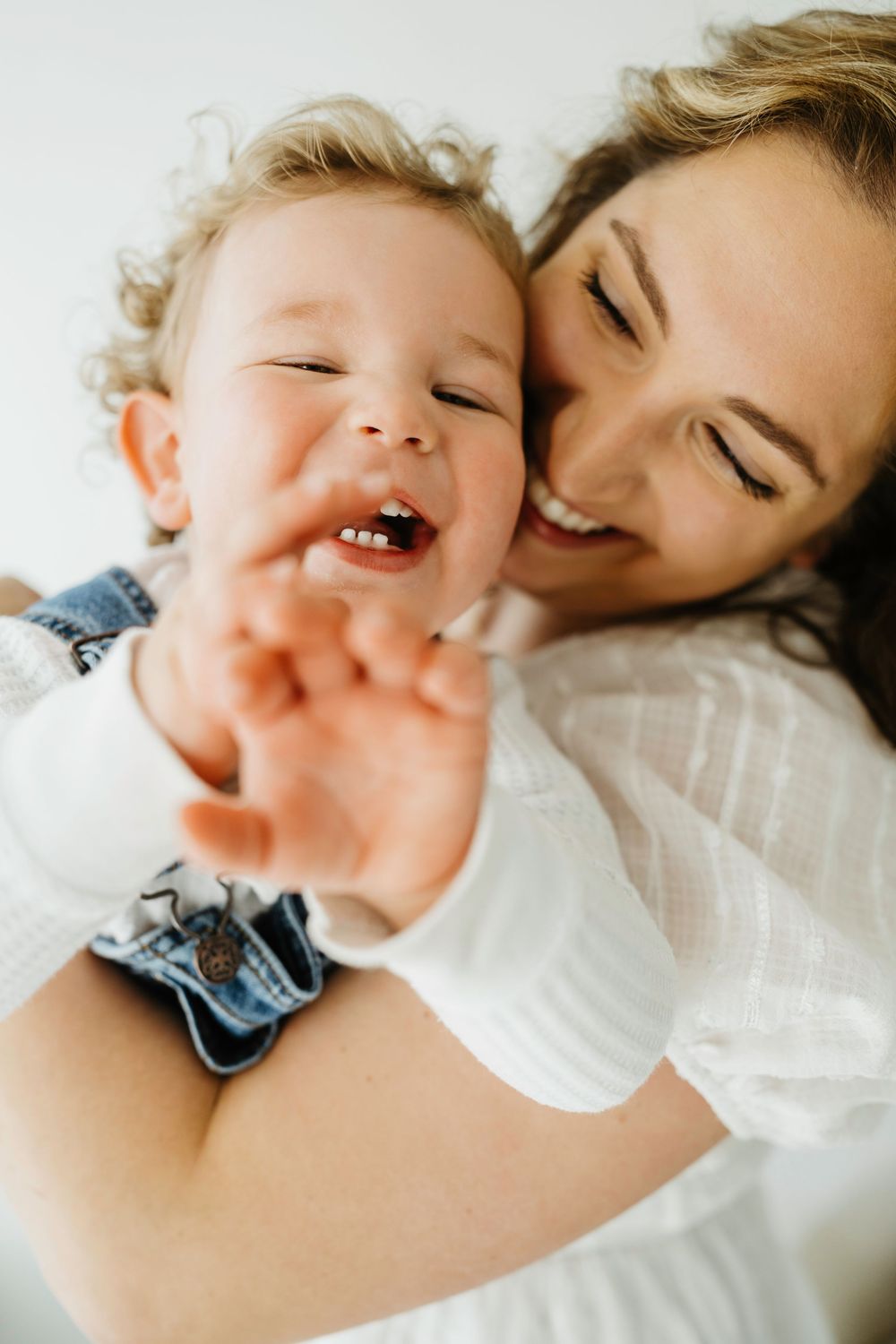 A joyful mother in white holds her laughing toddler who reaches toward the camera in a bright, cheerful moment.