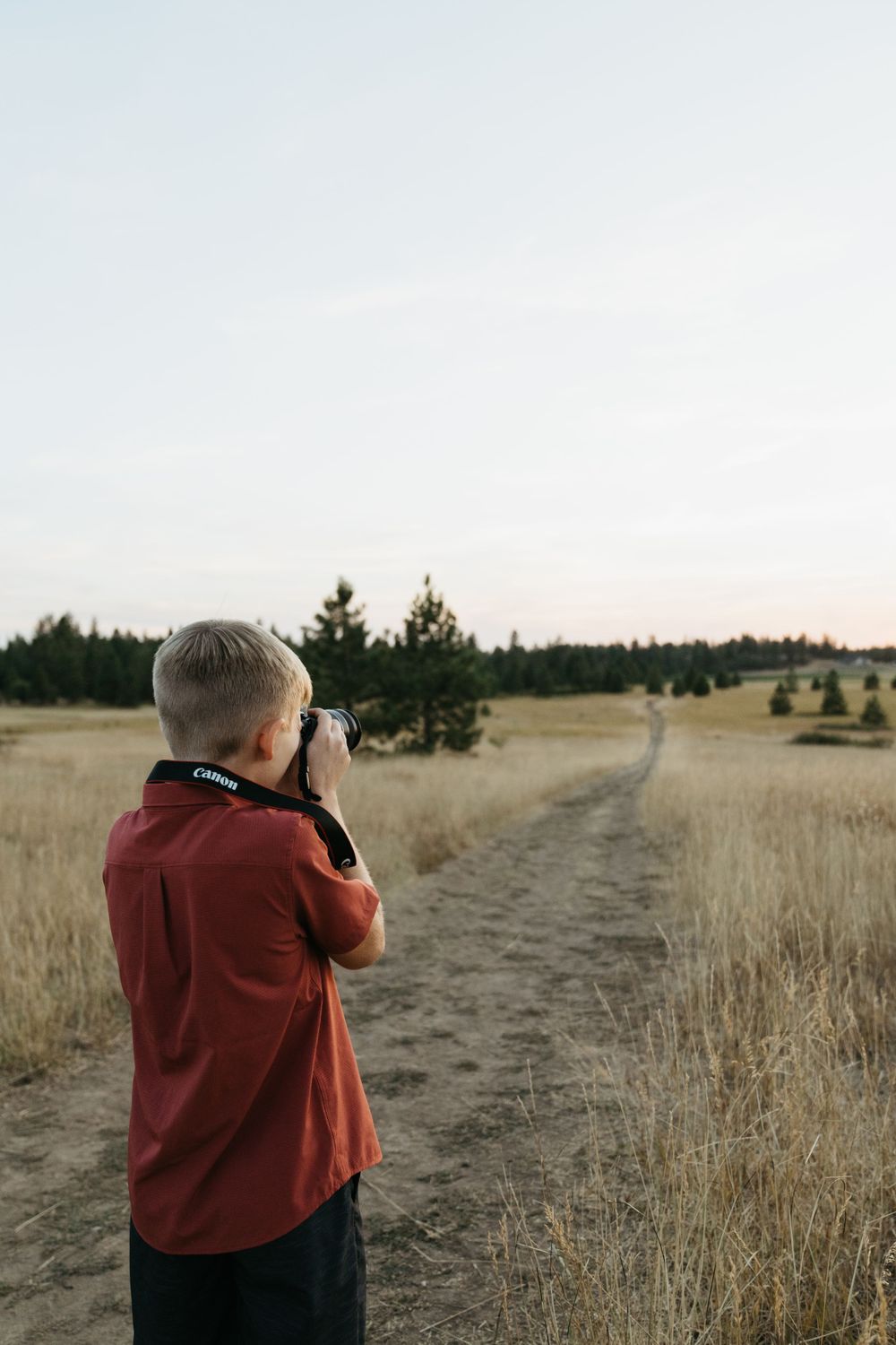 A photographer in a red shirt takes pictures on a dirt path through golden grasslands at sunset.