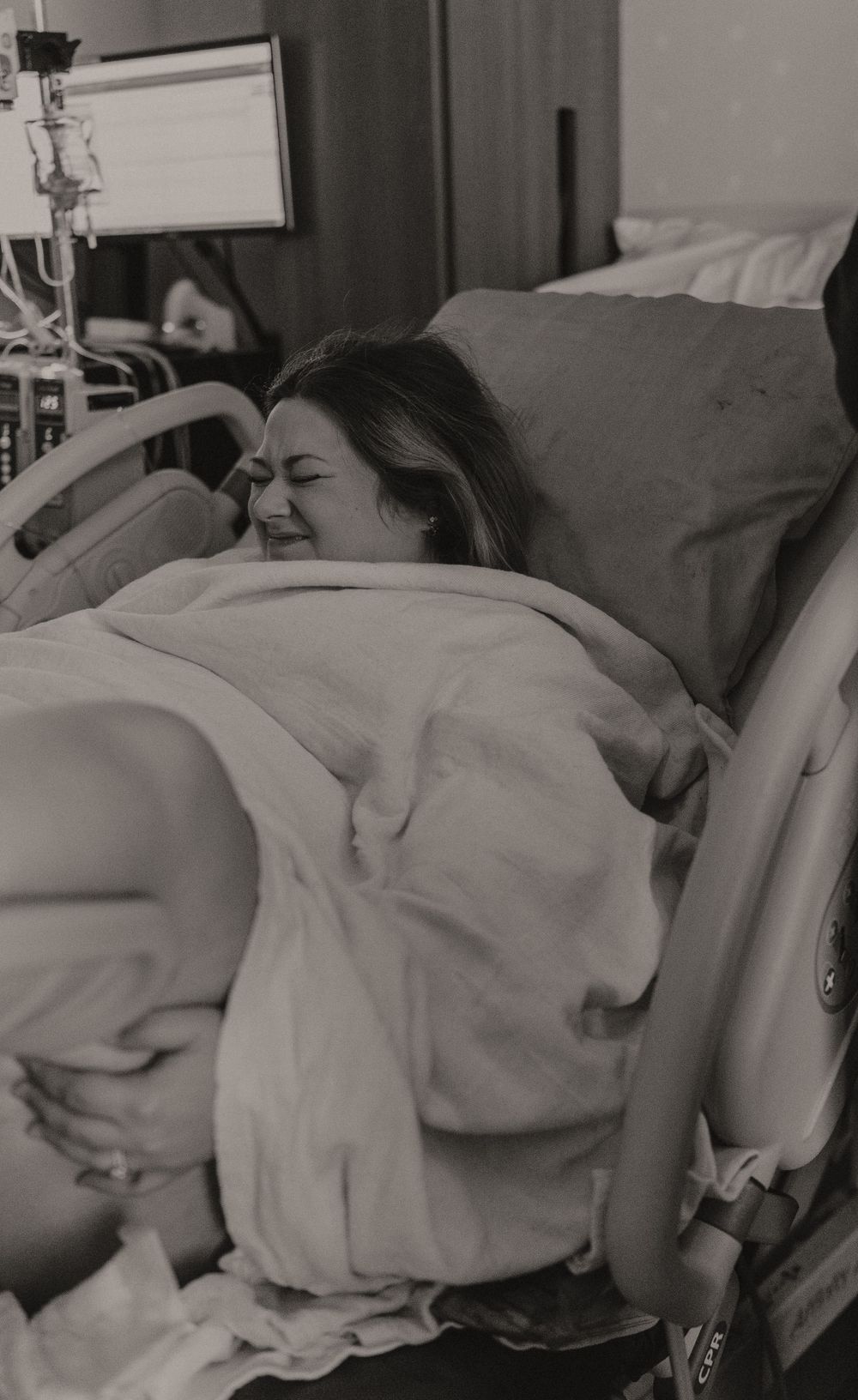 Hospital patient resting in bed under blankets with medical monitoring equipment visible.