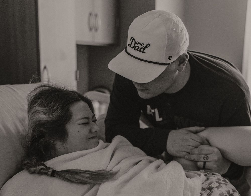 Hospital scene with patient resting while being comforted by visitor wearing baseball cap.