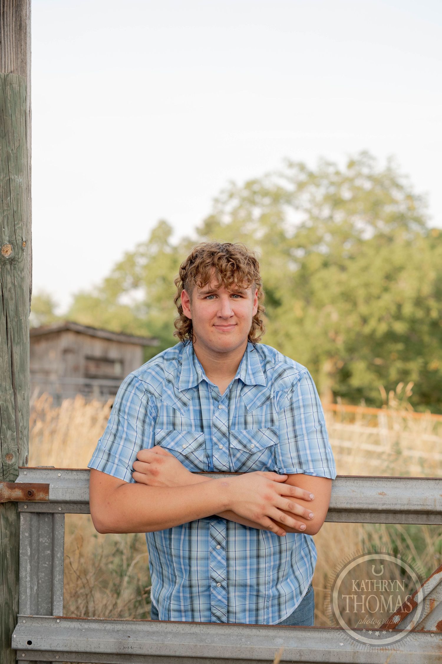 A casual portrait of a person in a blue plaid shirt leaning on a wooden railing in a rural setting.