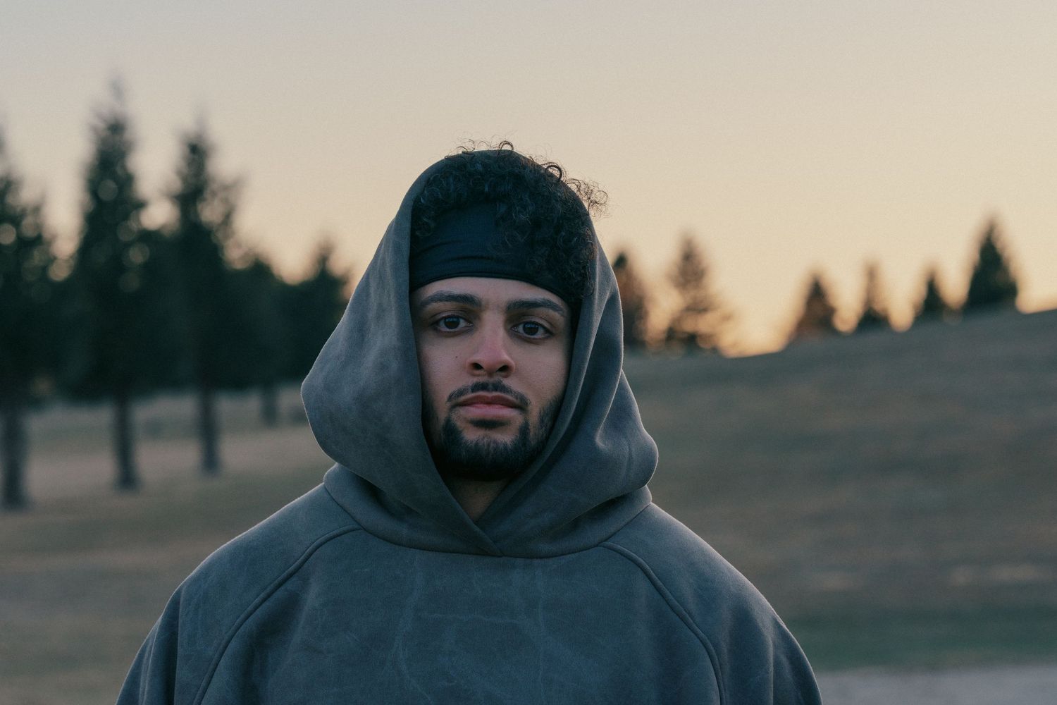 Person in grey hoodie standing outdoors at dusk with evergreen trees in background.