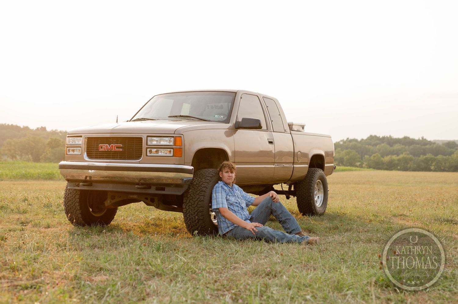 A lifted GMC pickup truck parked on grass with someone sitting against it during golden hour.