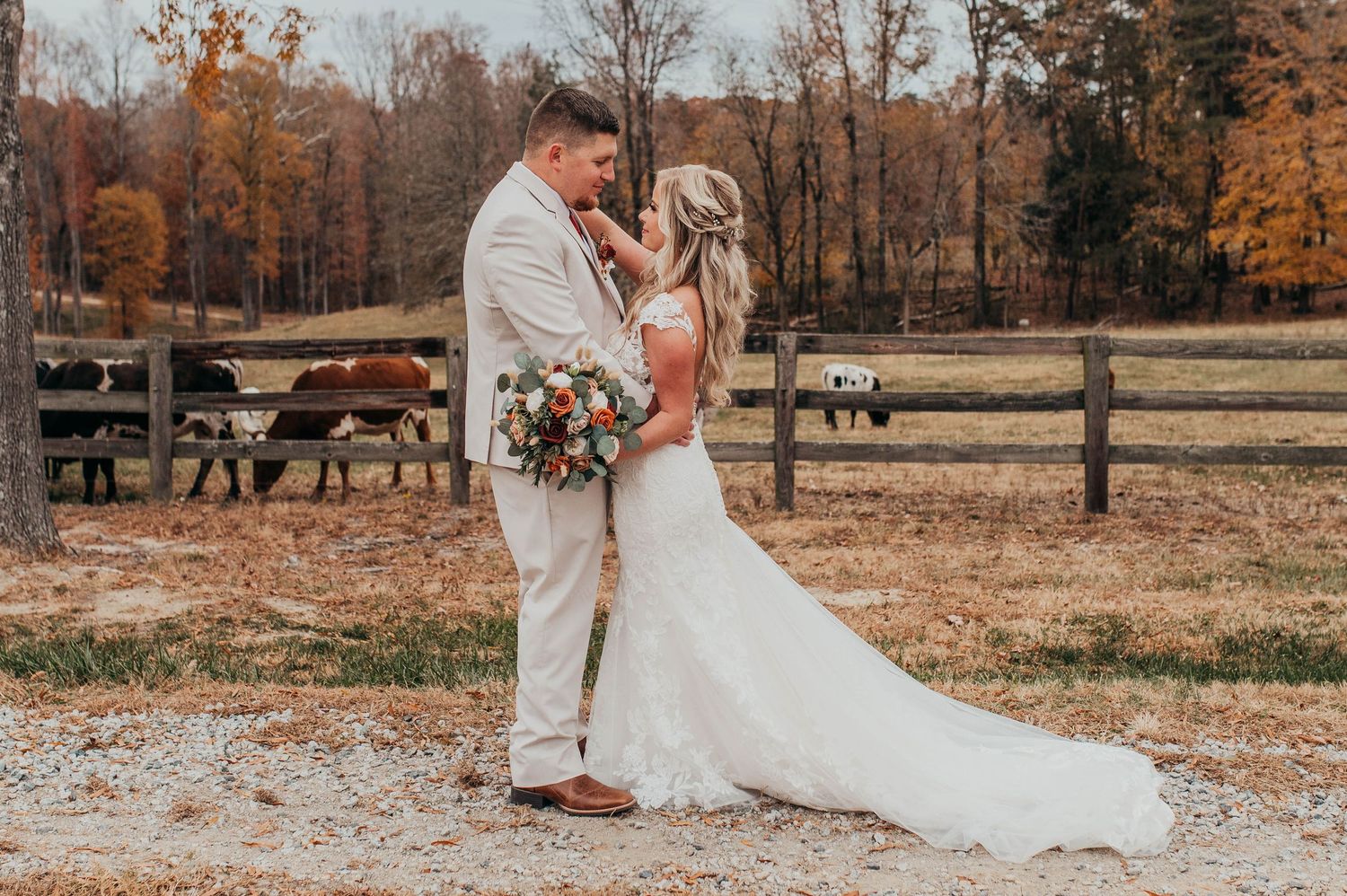 A couple embraces in their wedding attire on a fall day with wooden fences and autumn trees in the background.