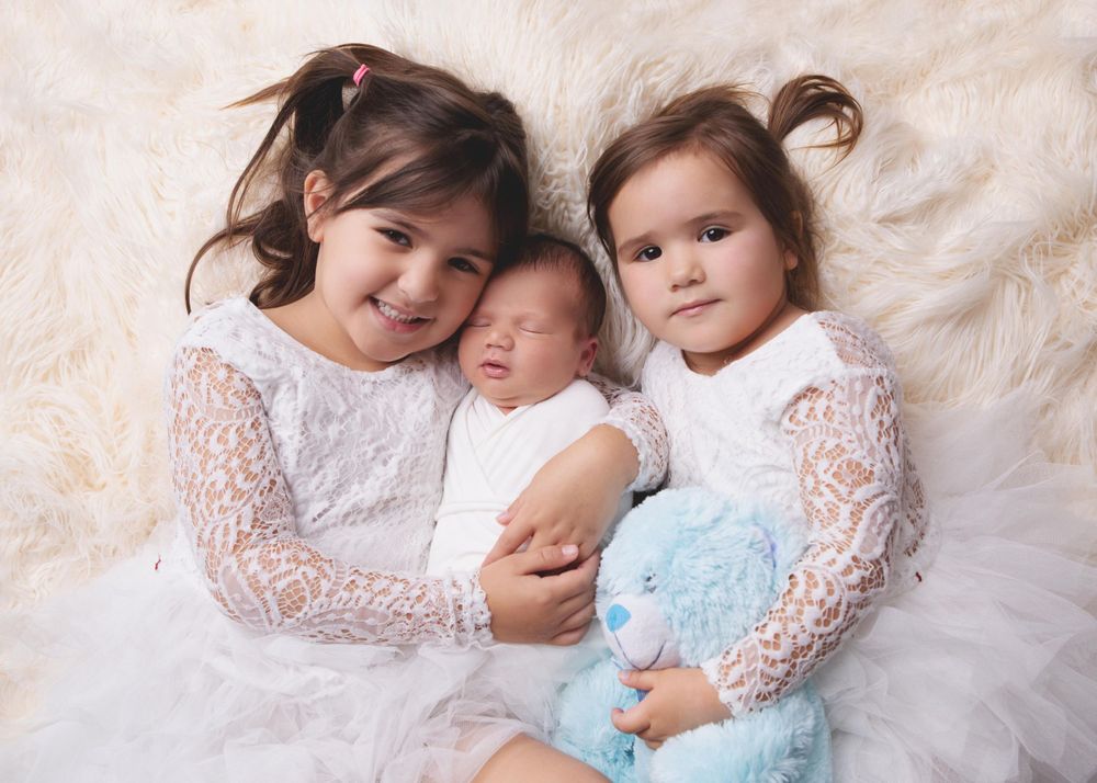 Three young children in white lace dresses pose together on a soft ivory fur backdrop.