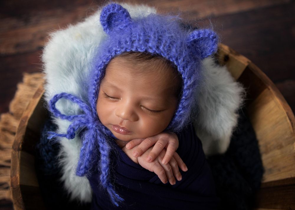 Sleeping baby wearing purple knit bear bonnet rests peacefully in wooden bowl with soft white fur lining.