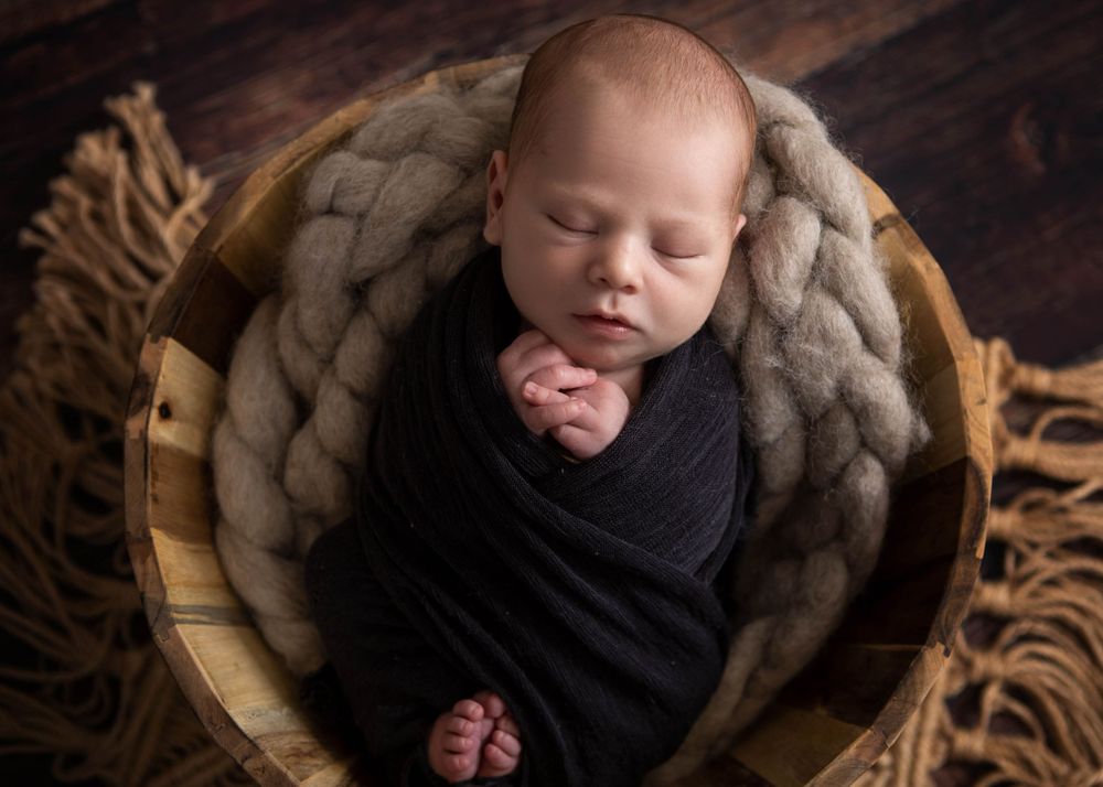 Sleeping newborn wrapped in black swaddle rests in wooden bowl with chunky knit blanket backdrop.