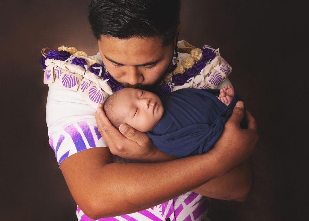 A tender embrace between parent and sleeping infant against a dark background with purple floral fabric.