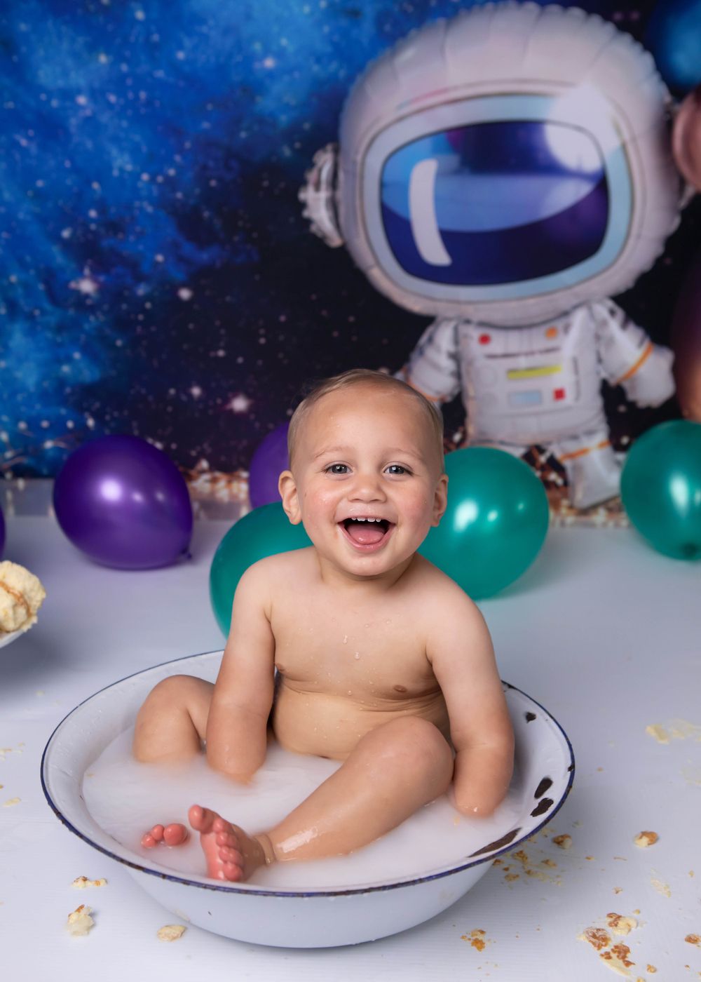Joyful baby sitting in a flying saucer prop with space-themed balloons and astronaut decorations in the background.