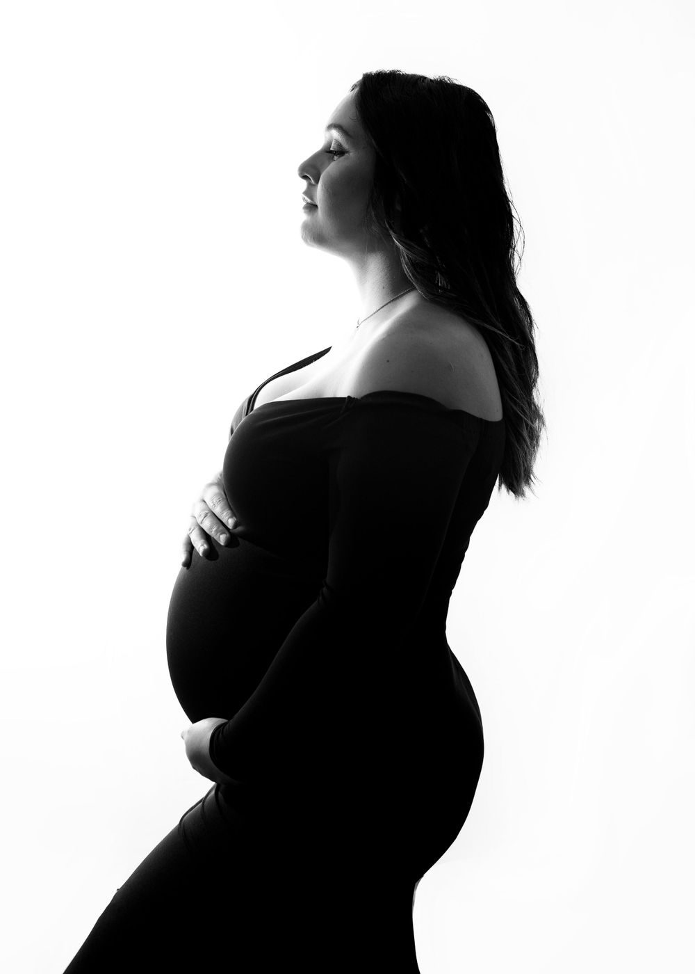 Black and white silhouette of a pregnant woman in profile wearing an off-shoulder dress against a white backdrop.