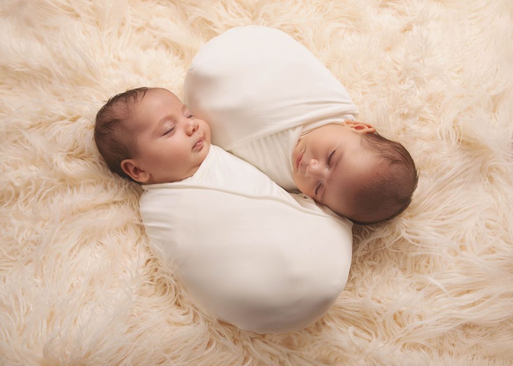Two sleeping newborn babies swaddled in white blankets lie together on a soft cream-colored fur blanket.
