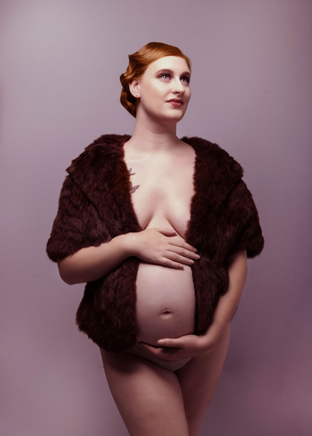Artistic portrait of an expectant mother wearing a dark brown fur wrap against a purple studio background.