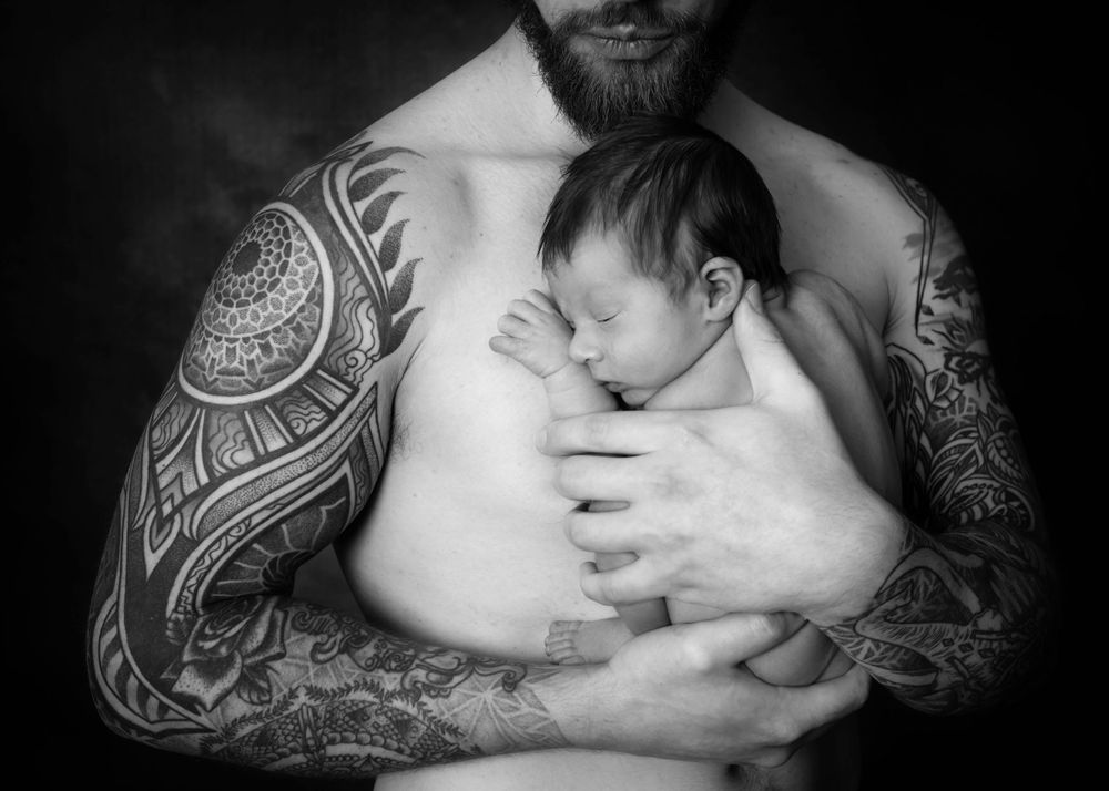 A black and white portrait of a newborn baby sleeping against a tattooed chest.