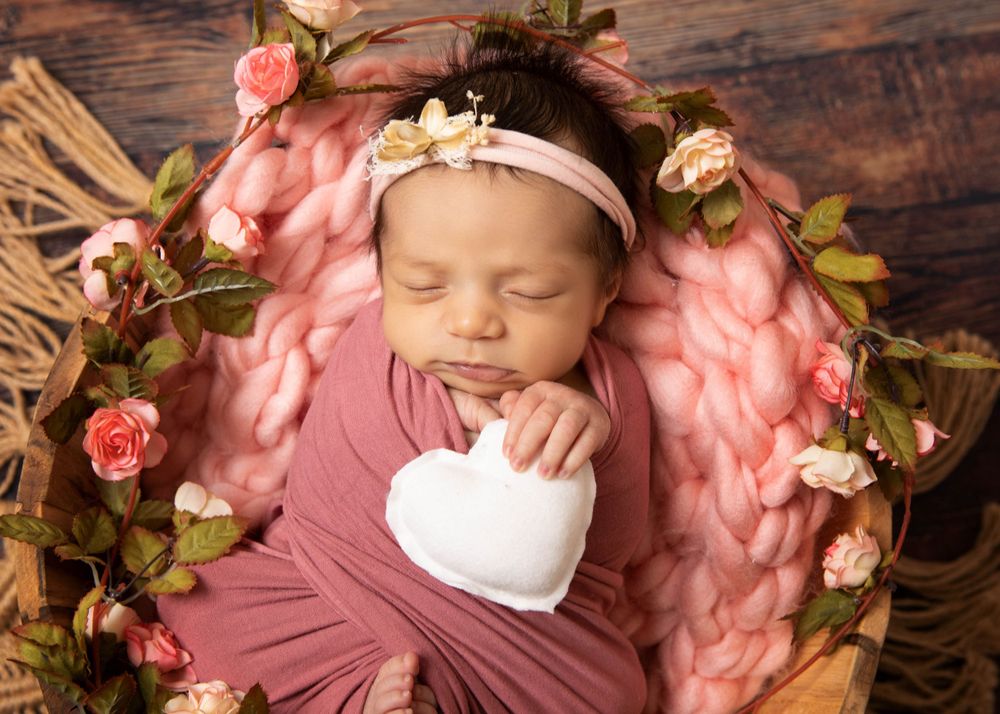 Newborn baby sleeping in wooden bowl with pink floral decorations and delicate headband.