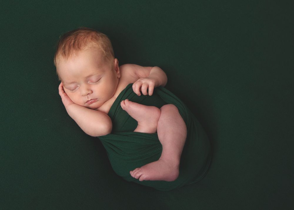 Peaceful newborn baby sleeping wrapped in dark green fabric against matching background.