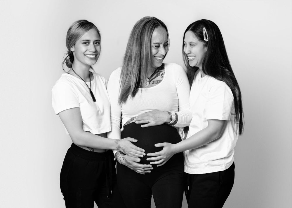 Three friends in white shirts celebrating a pregnancy with hands on belly in black and white photo.
