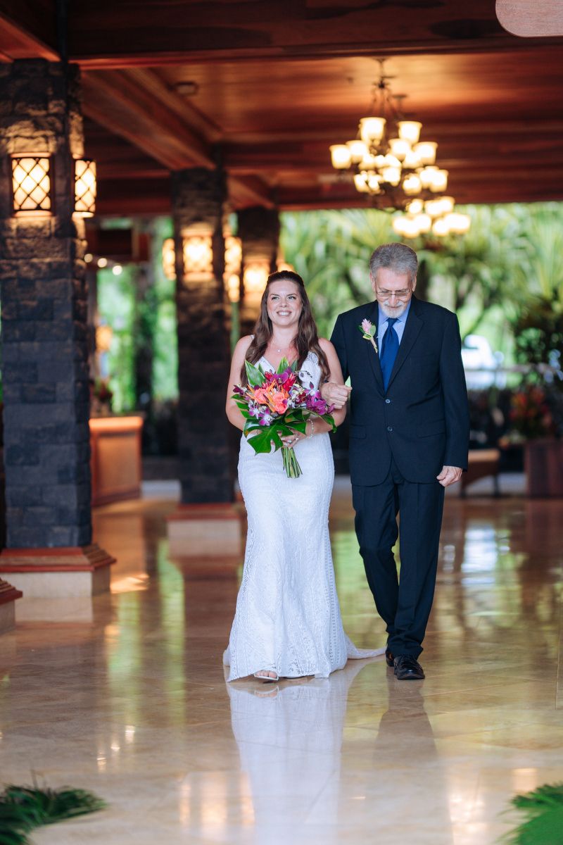 A bride in a white lace gown walks down an elegant corridor with chandeliers and stone pillars while holding a vibrant bouquet.
