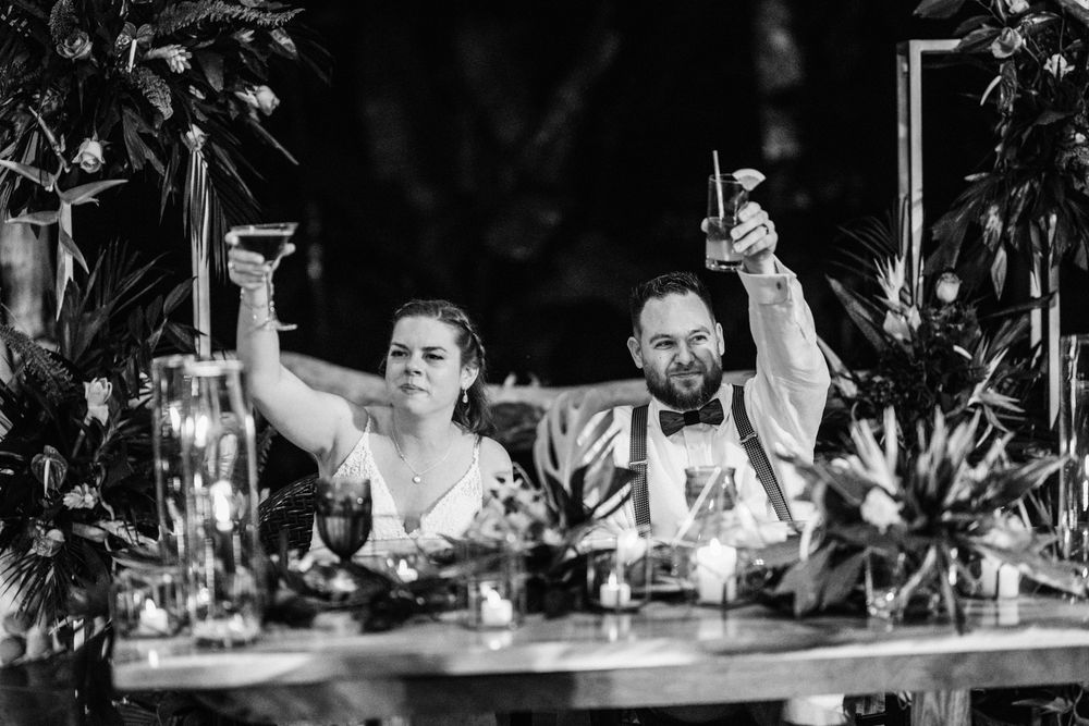A black and white photo of a couple raising their glasses in a toast at an outdoor reception table with floral decor.