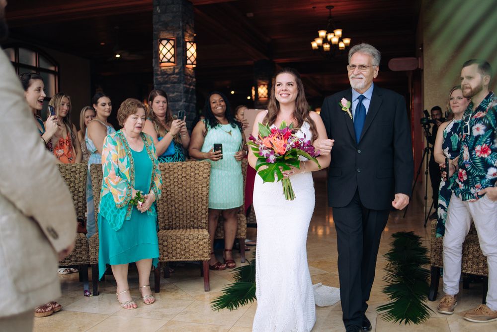 A wedding ceremony takes place in an elegant indoor venue with guests watching as someone in white walks down the aisle.