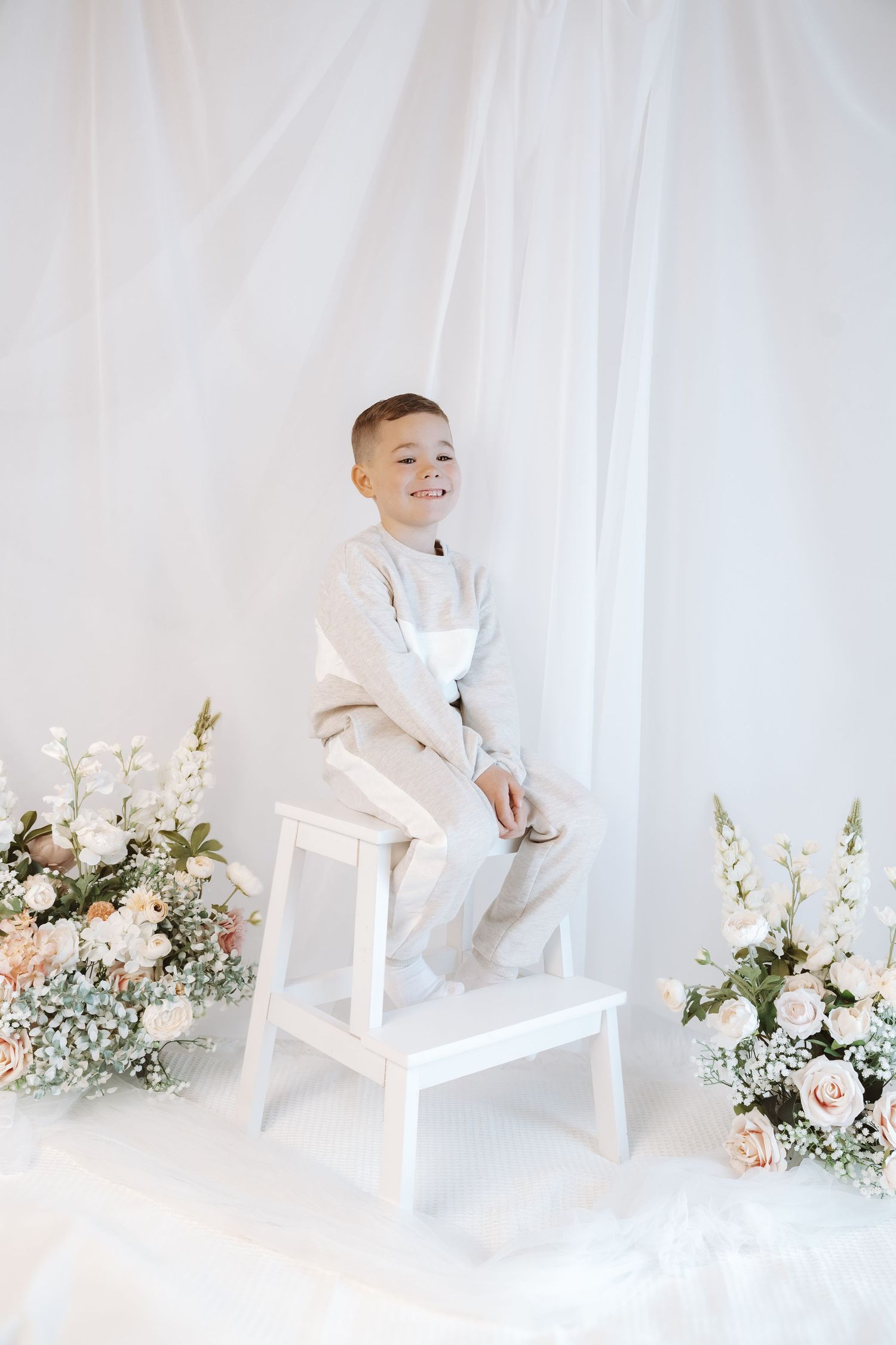 Young child in all white formal attire sits on white step stool surrounded by elegant floral arrangements.