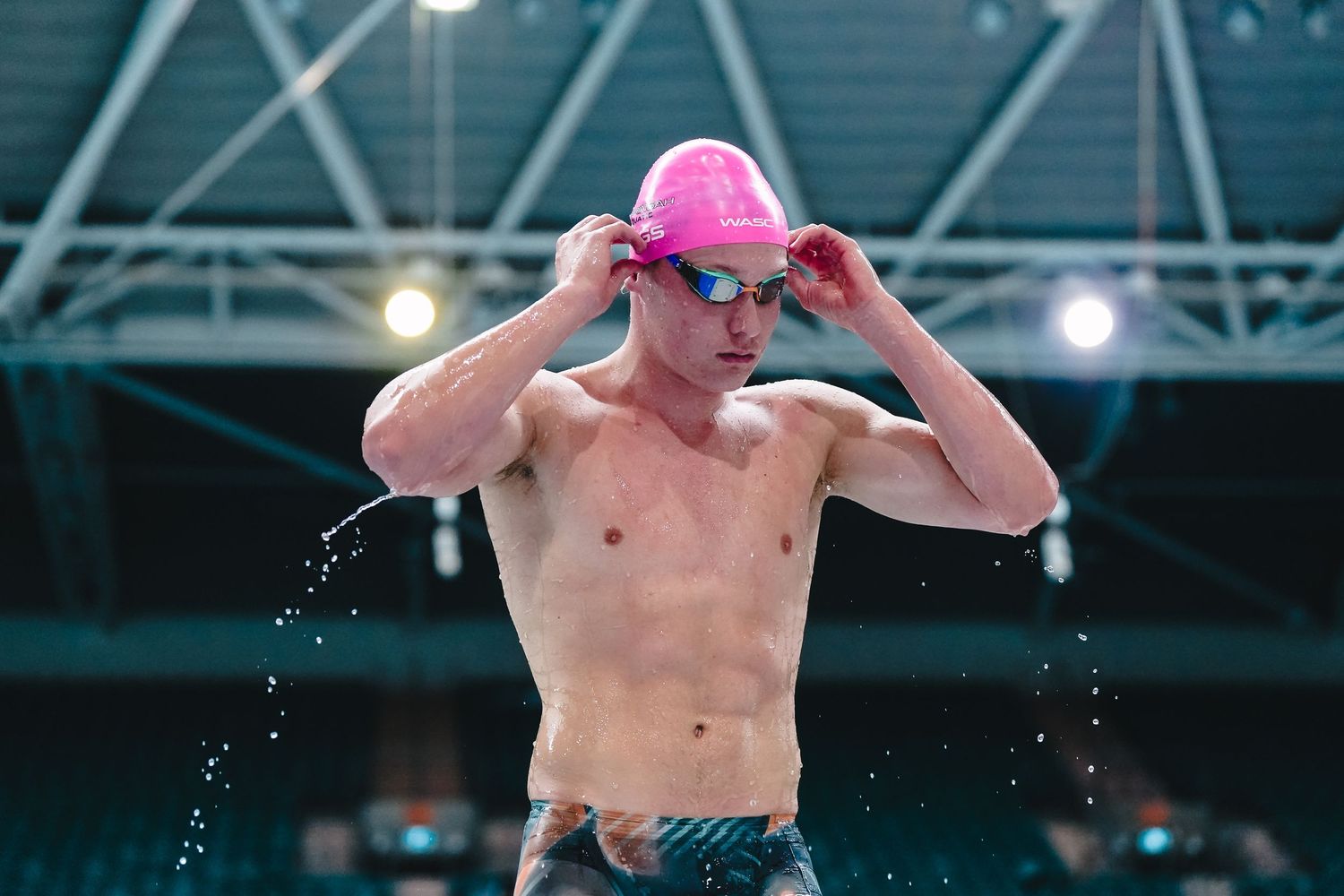 Competitive swimmer adjusts pink swim cap at indoor pool with water droplets flying and arena lights overhead.