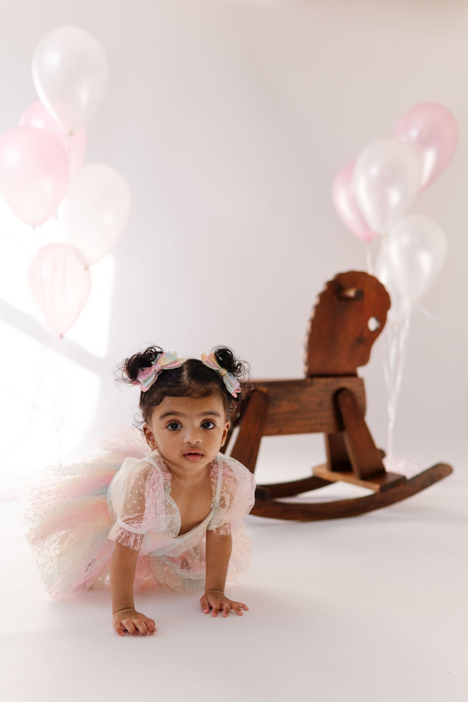 Baby in white lace dress crawls near wooden rocking horse with pink and white balloons in background.