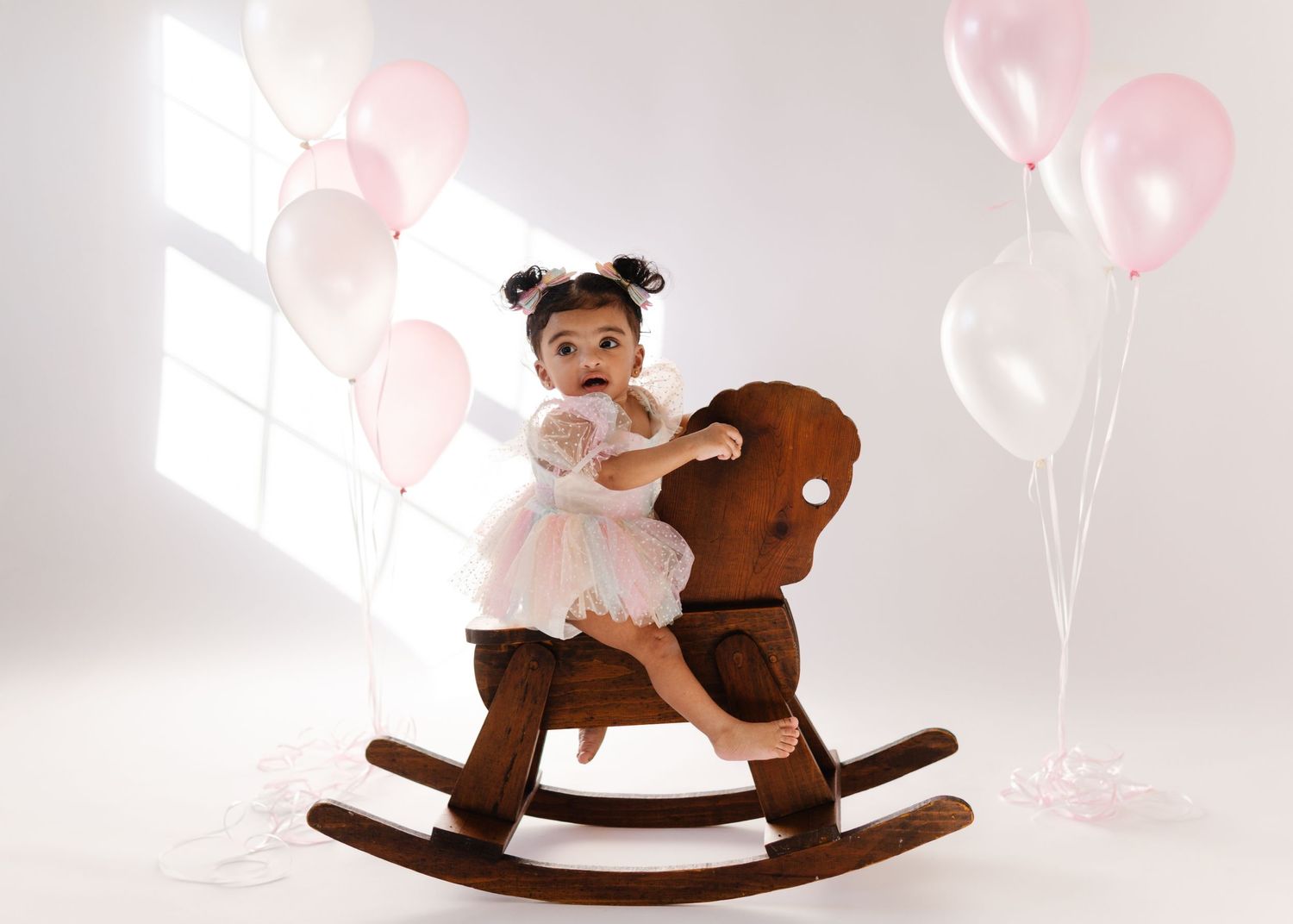 A toddler in a white dress sits on a brown wooden rocking horse surrounded by pink balloons in a bright room.