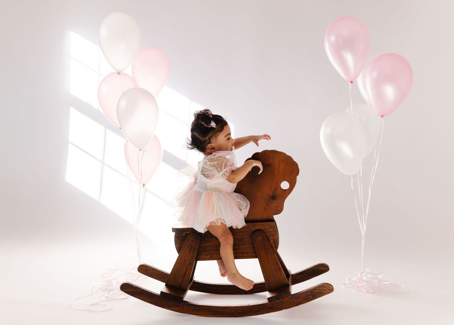 Child in white dress plays on wooden rocking horse surrounded by pink balloons in bright studio setting.