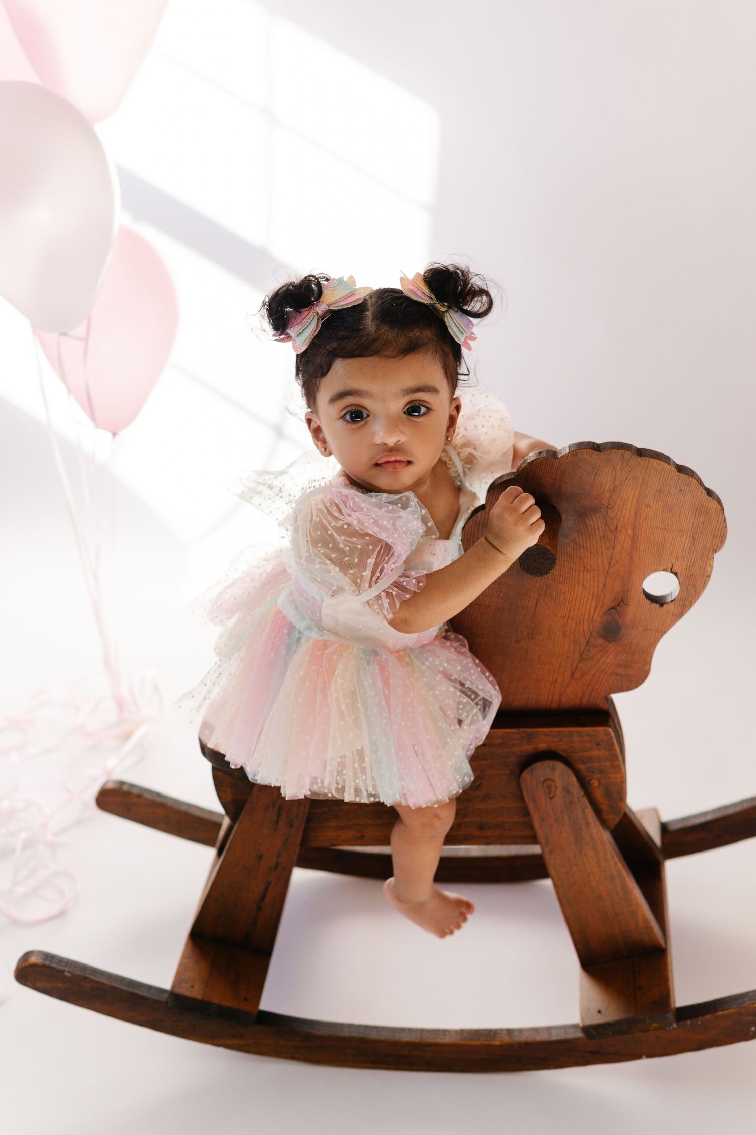 A small child in a white tulle dress balances on a wooden rocking horse surrounded by pink balloons.
