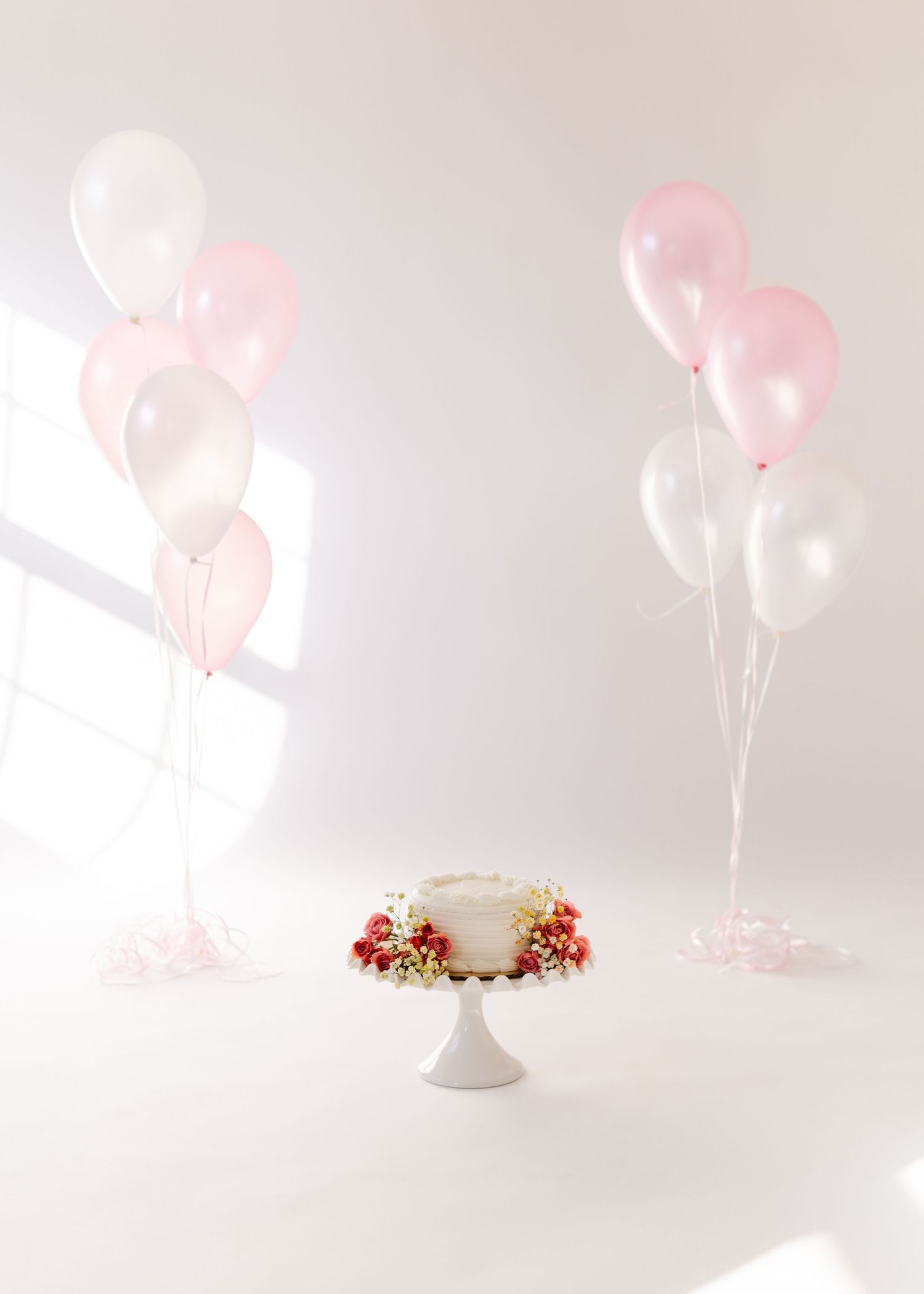 A white cake on a pedestal stand decorated with red flowers, surrounded by pink and white balloons against a light backdrop.