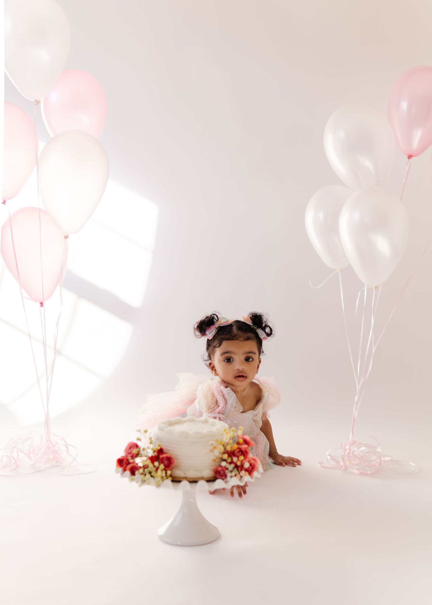 First birthday cake smash photo with pink balloons and white cake on pedestal against light background.