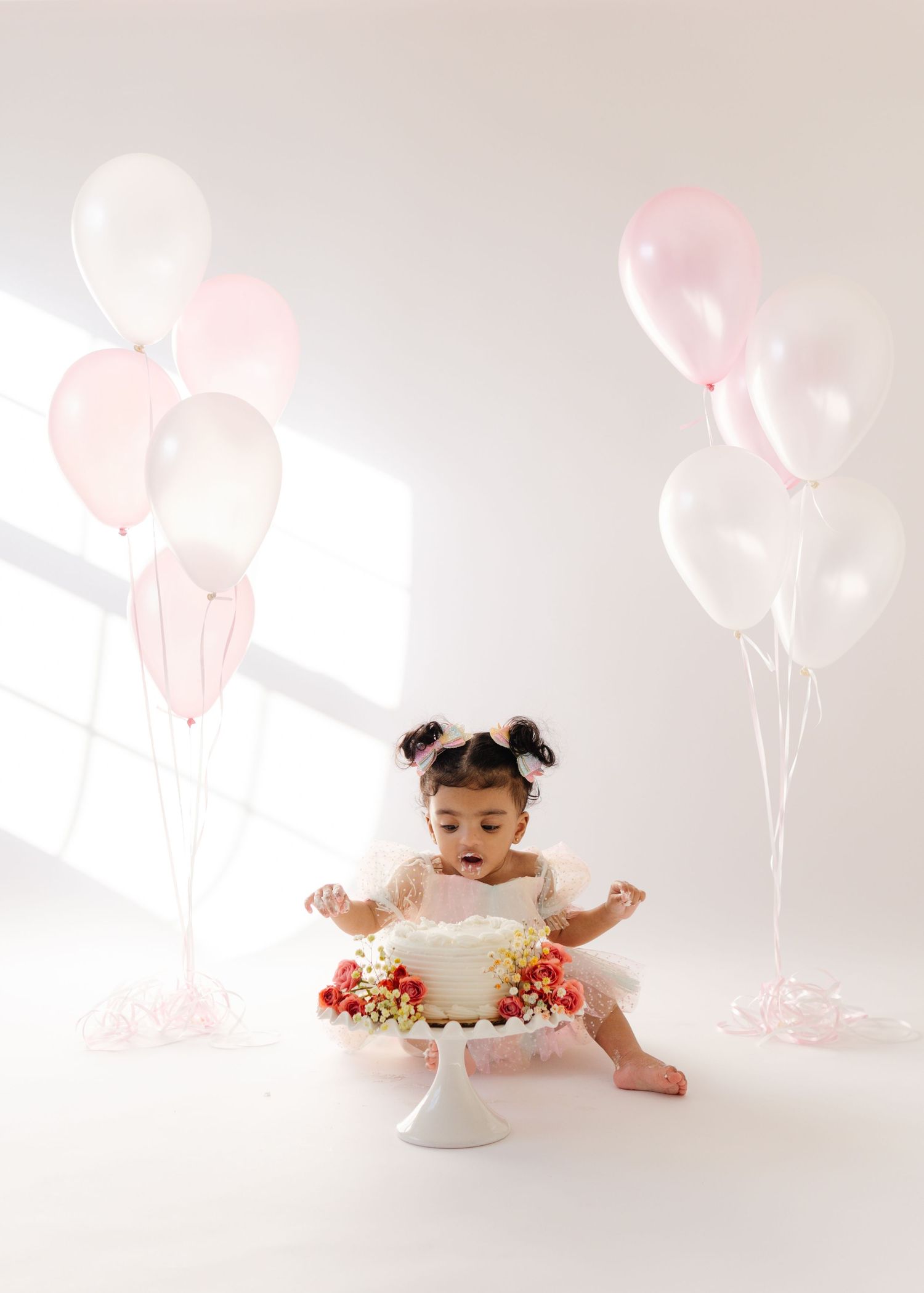 Baby in white dress seated on cake stand with pink balloons in studio setting for first birthday cake smash photo shoot.