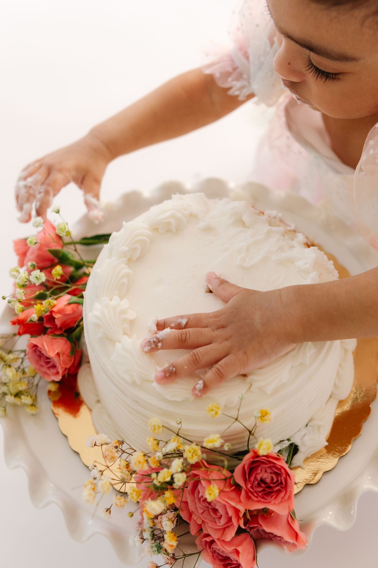 Fresh white cake decorated with coral roses and floral accents being carefully adorned by hands from above.