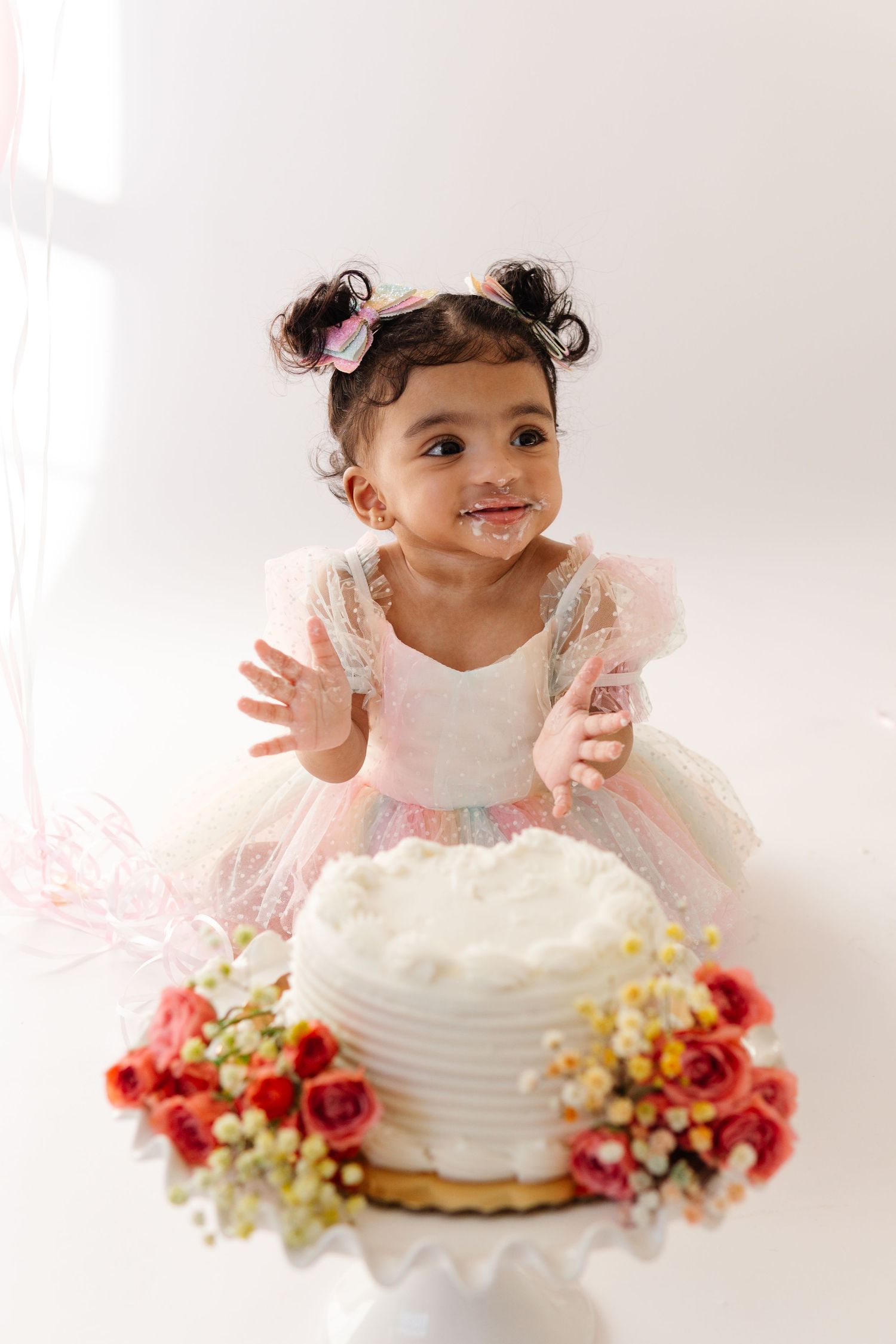A small child in a pink tulle dress leans over a white frosted cake decorated with vibrant red and pink flowers.