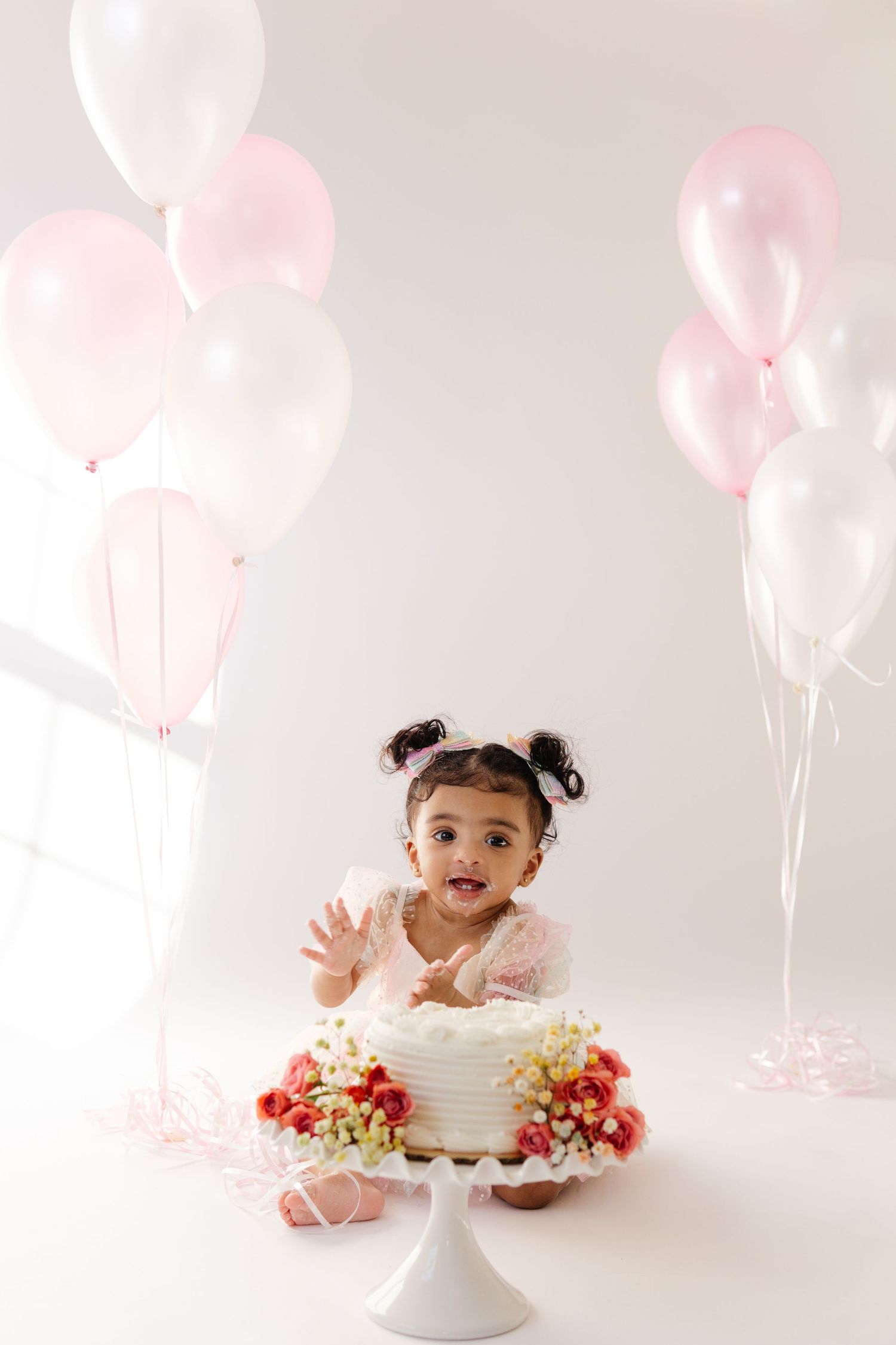 Pink and white balloons decorate a simple cake smash photo session with a white cake on a pedestal stand.