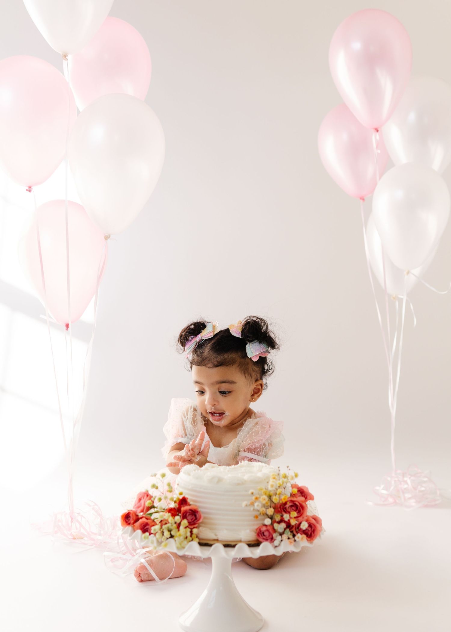 A small child sits by a beautiful white cake decorated with flowers and pink balloons in the background.